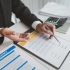 A businessman in a formal suit explains car insurance details, holding a small red car model and pointing at contract documents, symbolizing agreement, protection, and financial planning.
